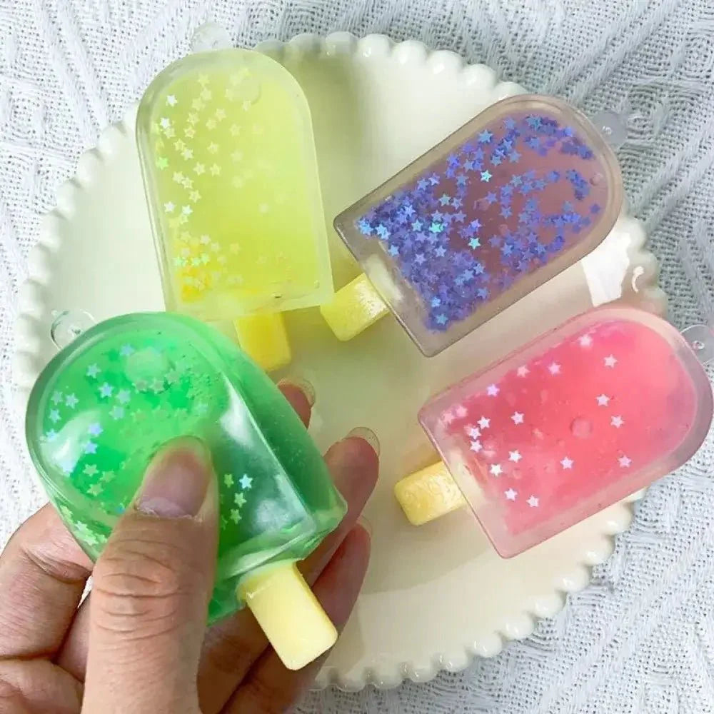 Une main tient un squishy creme glacee vert translucide en gel avec paillettes étoiles blanches parmi des bonbon ourson colorés