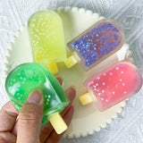 Une main tient un squishy creme glacee vert translucide en gel avec paillettes étoiles blanches parmi des bonbon ourson colorés